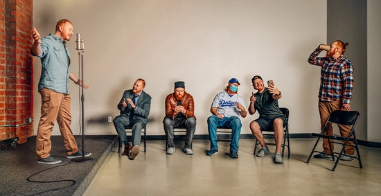 Thomas Hagena stands near a microphone while 5 clones of himself stand/sit watching, all wearing different clothes and showing slightly different personalities.