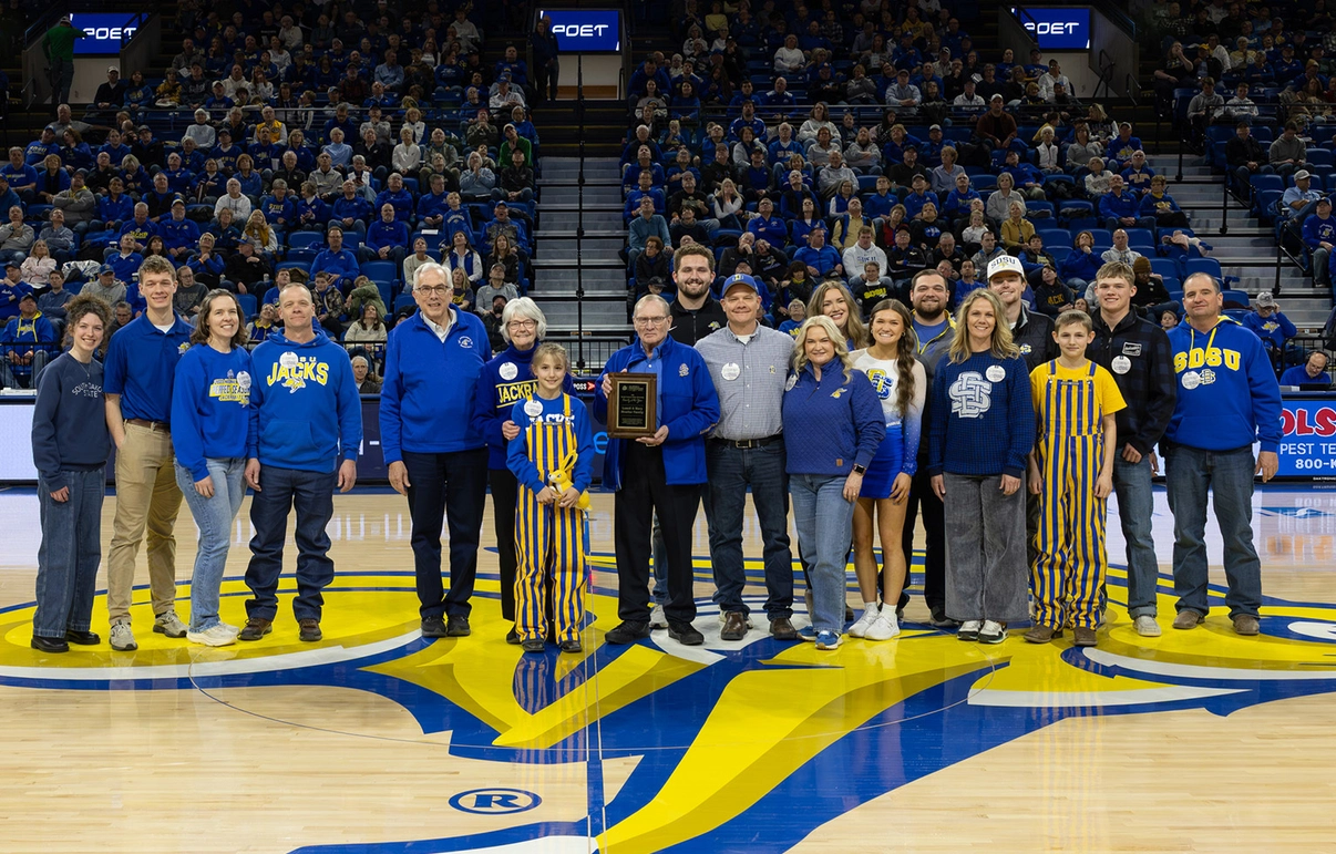 A large group of the Moeller Family, all wearing Jackrabbit gear, stands together at half court of First Bank & Trust Arena with President Barry Dunn.