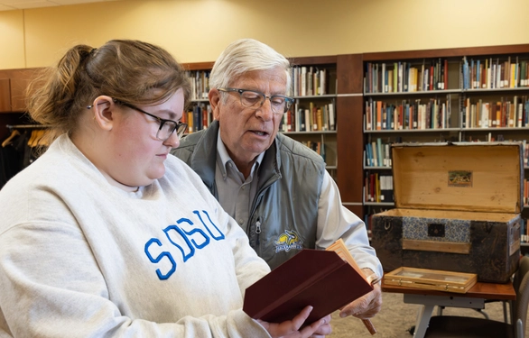 Two people examine a book in a library.