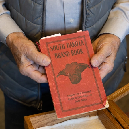 A person holds a red book titled "South Dakota Brand Book." The cover features a cattle silhouette.
