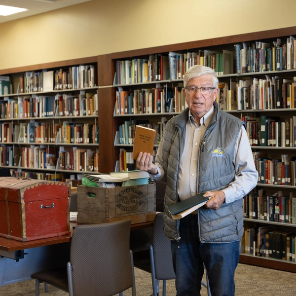 Myron Sonne stands in a library holding a book, surrounded by shelves filled with books.