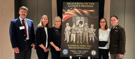 Five people stand beside a poster for National POW/MIA Recognition Day, displaying military figures and an American flag, conveying respect and commemoration.
