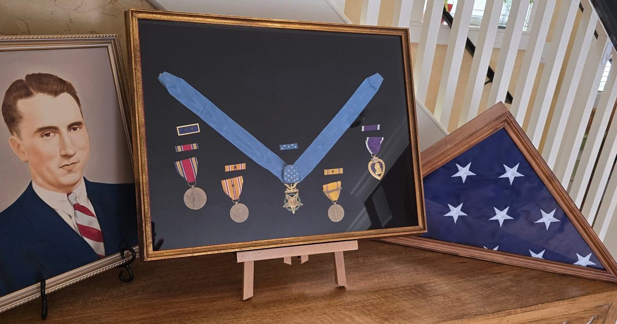 Portrait of a Willibald Bianchi, military medals in a display case, and a folded American flag on a wooden table beside a staircase.