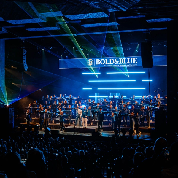 A large group of singers performs on a dimly lit stage with blue neon lights and "Bold & Blue" sign above.