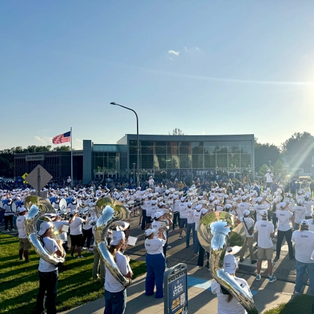 A large marching band in white uniforms perform outside the SDSU Alumni Center in bright sunlight.