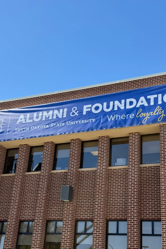 A worker on a blue lift hangs a large blue banner reading "SDSU Alumni & Foundation" on a brick building under a clear blue sky.