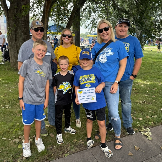 A group of seven people, including adults and children, stands on a grassy area under trees by the SDSU Alumni & Foundation's tailgate tent.