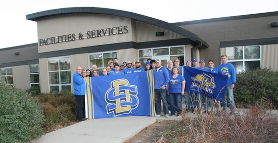 A group of people in blue and yellow shirts holding a large blue and yellow SDSU banner outside of the Facilities & Services building.