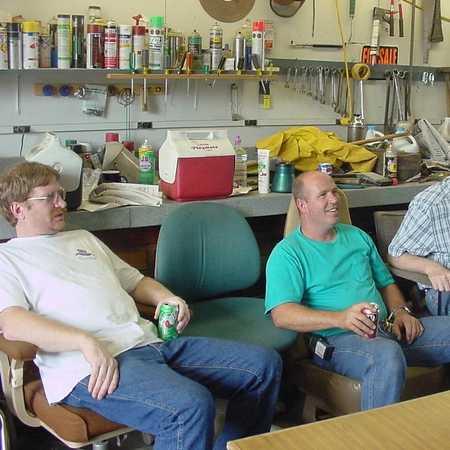 A photo of four men sitting in a workshop with tools and supplies behind them.