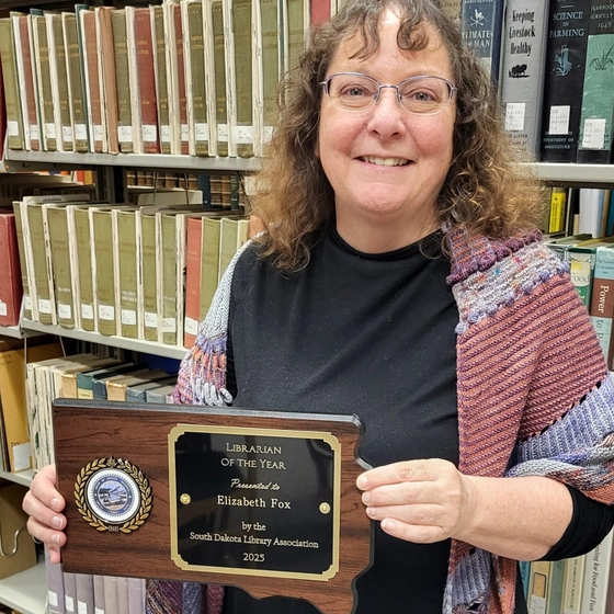 Elizabeth Fox smiling and holding her Librarian of the Year wooden plaque in a library.