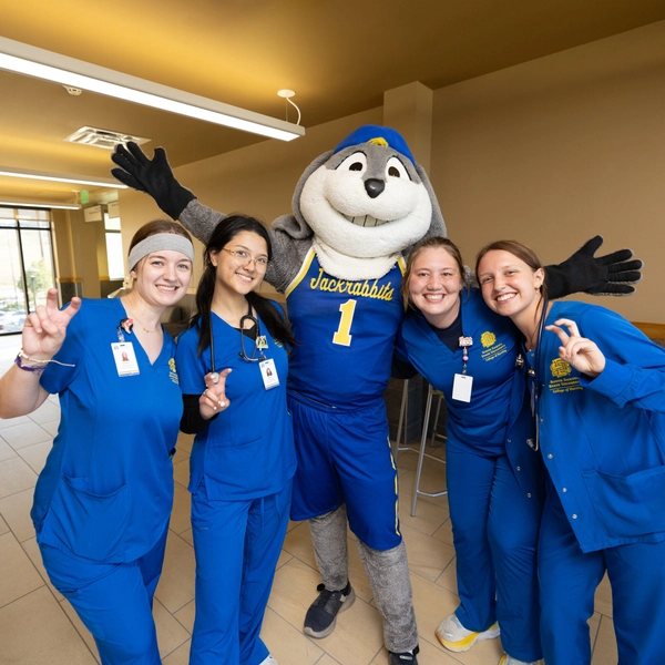 A group of four nursing students wearing blue scrubs and the SDSU mascot Jack stand for a photo looking cheerful.