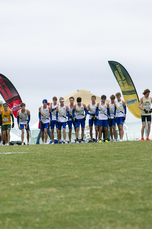 Athletes in colorful uniforms line up at the start of a race on a grassy field, with flags behind them and a white tent in the background under an overcast sky.
