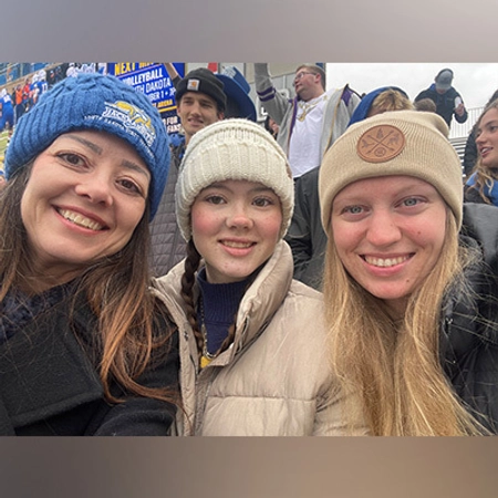 Three people in SDSU winter attire, smiling and posing for a selfie at a football game.