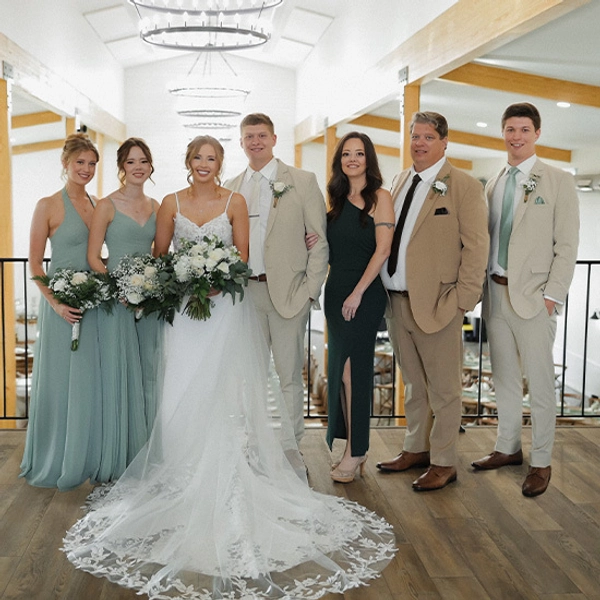Wedding party posing indoors. The bride in a white gown stands with bridesmaids in teal dresses and groomsmen in beige suits, smiling warmly.