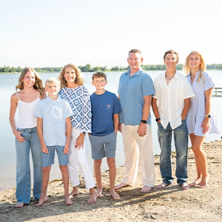 A family of seven stands barefoot on a beach, smiling under a clear sky.