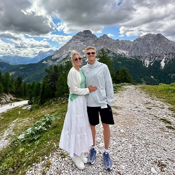 A couple stands on a rocky path with picturesque mountains and greenery in the background.