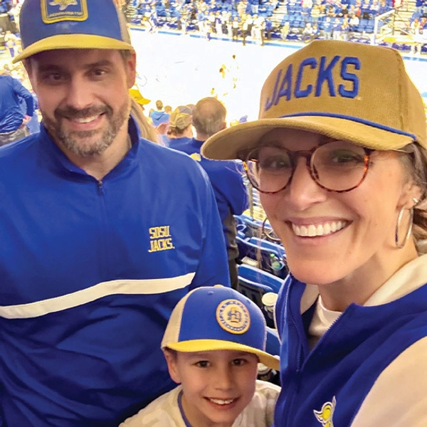 Megan DeBoer with her partner, Anthony, and her son, smiling for a photo at a Jacks basketball game.