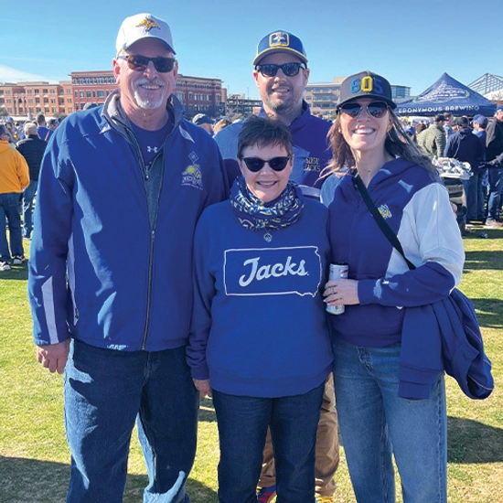 A group of four people smiling outdoors at a sports event, wearing South Dakota State University apparel.