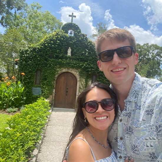 Nick and his wife are smiling towards the camera in front of a church covered in greenery.