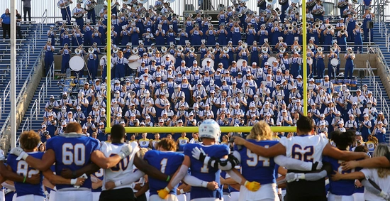 SDSU Football team stands next to one another singing school song after a game, with The Pride of the Dakotas in the background playing.