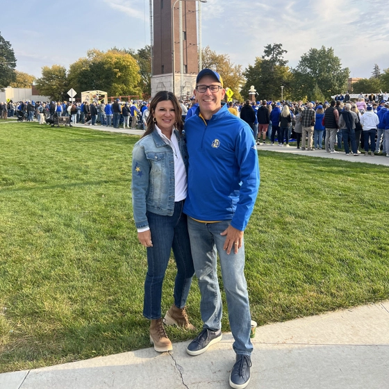 Tracy and Wes Chambers are smiling and standing on the grass outside the SDSU Alumni Center with the Coughlin Campanile behind them.