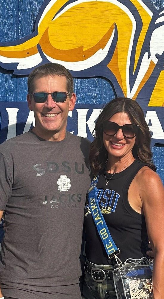 Wes and Tracy Chambers are smiling and standing in front of a blue wall with a large Jackrabbit logo and the word "Jackrabbits" in text.