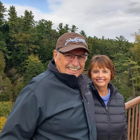 Gary and Donna Dettman are standing on a bridge surrounded by lush green trees.
