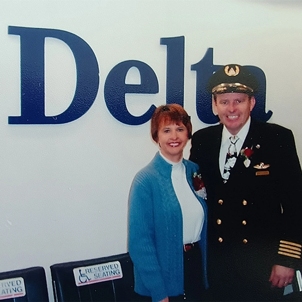 Donna and Gary Dettman standing in front of a Delta Airlines logo. Gary is wearing a pilot's uniform and Donna is wearing a blue sweater.