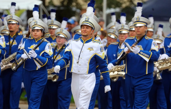Evan Bartelt, wearing a white uniform, is leading the marching in front of The Pride of the Dakotas Marching Band, who are wearing blue uniforms.
