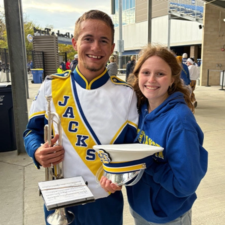 Smiling Evan Bartelt, wearing a white-and-blue Pride of the Dakotas uniform, holds a trumpet beside a woman in a blue hoodie at an outdoor event.