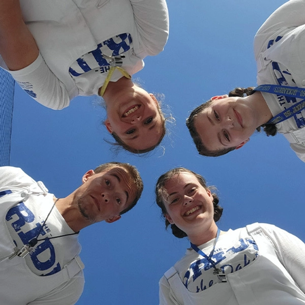 The four majors of the Pride of the Dakotas Marching Band, smiling in white T-shirts, looking down at the camera against a blue sky.