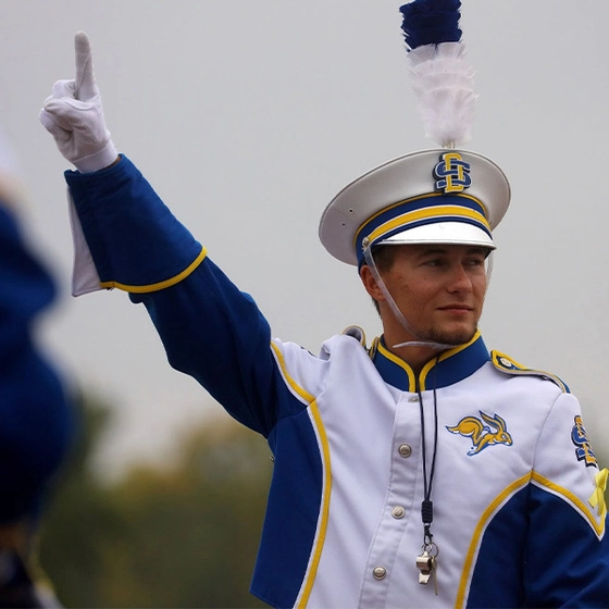 Evan Bartelt, in a white, blue, and yellow Pride of the Dakotas marching band uniform, raises a gloved finger to the band while conducting.