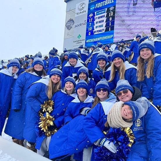 Hannah Hank and a group of other members of the Pride of the Dakotas smile together in a snow-packed stadium, all wearing long blue winter coats.