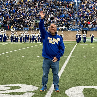 Jeff Eckhoff in a blue SDSU sweater waving to a cheering crowd from the 20-yard line on the football field at Dana J. Dykhouse Stadium.