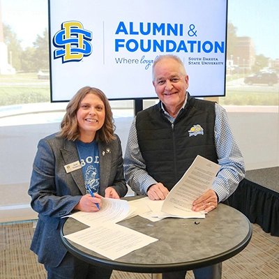 Andi Fouberg and Steve Erpenbach smiling for the camera while signing consolidation paperwork at a round table with the new SDSU Alumni & Foundation logo behind them.