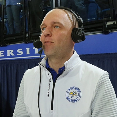 Tyler Merriam is wearing headphones and live-commentating at a sports event while wearing a white SDSU quarter-zip jacket.
