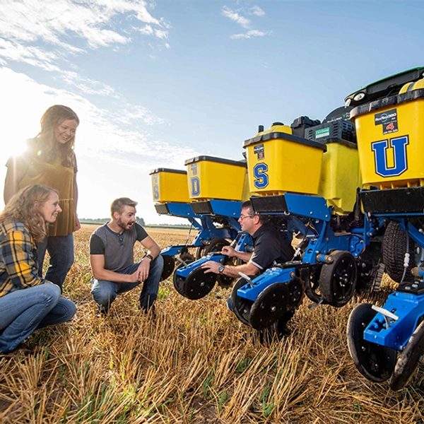students kneeling in a field with a professor with machinery in the background with decals on it that spell SDSU