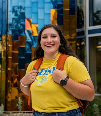Female SDSU student wearing a backpack, smiling and standing in front of the colored glass outside the American Indian Student Center.