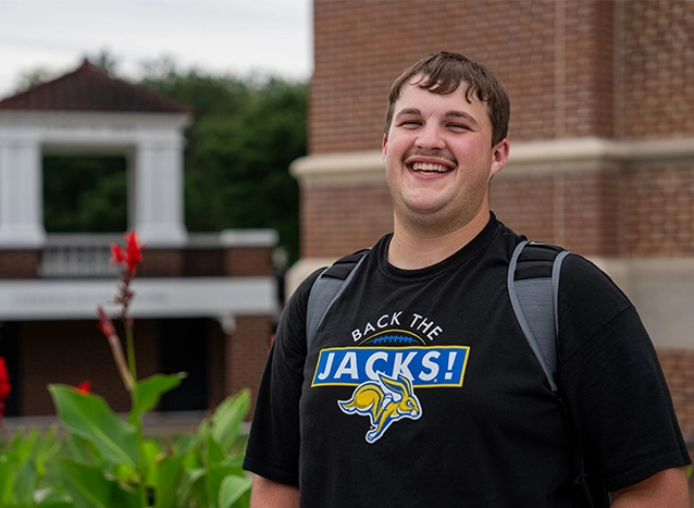 Male SDSU student wearing a backpack, smiling and standing outside Lincoln Hall.