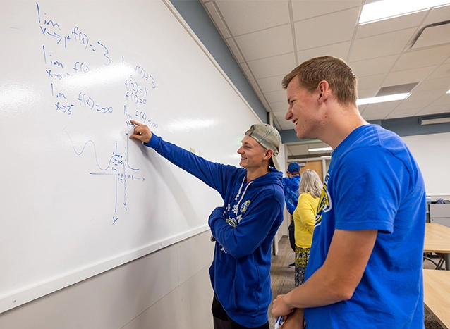 Two male students at a white board pointing at a math problem