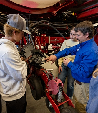 Male professor points and shows two students something on a piece of red farming equipment.