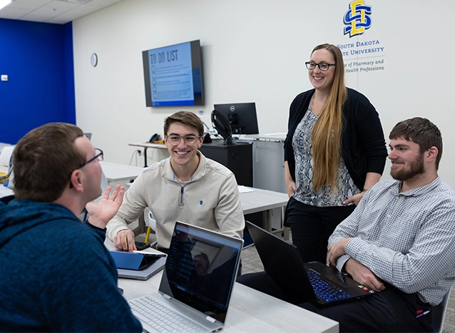 Female professor stands while talking with three male students sitting at a table with their laptops open.