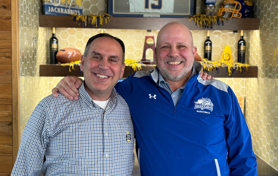 Kevin Kessler and Justin Sell stand together, smiling with SDSU memorabilia displayed on the shelves behind them.